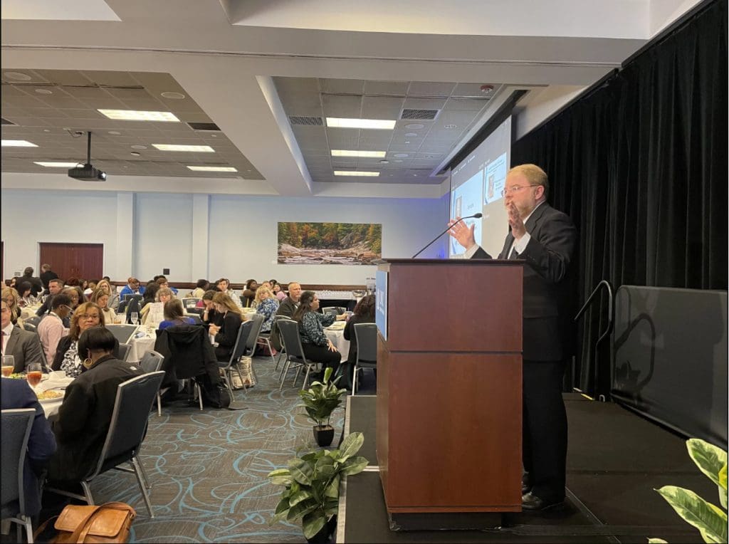 UNC System President Peter Hans speaks from a podium to a crowd of people seated at round tables at the 2023 Behavioral Health Convening