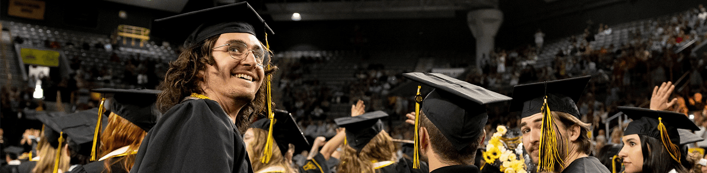 A graduation ceremony showing graduates in caps and gowns seated in an arena with a crowd in the background. One graduate in the foreground is smiling and looking upward.