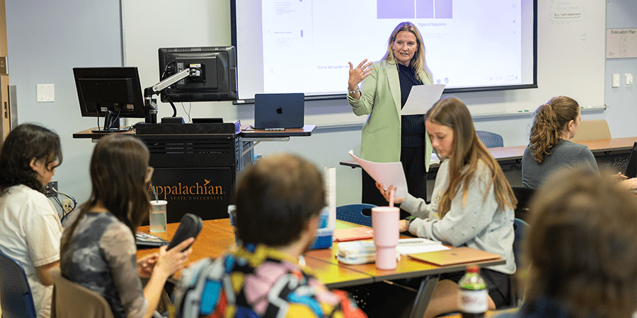 A female professor wearing a green blazer addressing college students seated at tables in a classroom.