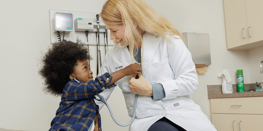 A young child using a stethoscope to listen to a female doctor's chest in a medical examination room. The scene highlights a playful interaction between patient and healthcare provider.