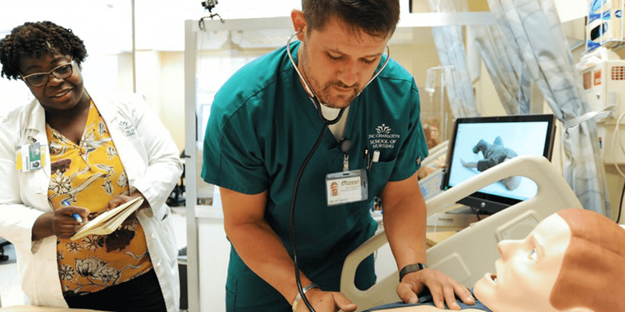 A medical training scenario showing a healthcare professional in green scrubs using a stethoscope on a medical mannequin lying in a hospital bed.