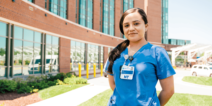 A nursing student wearing blue scrubs and an ID badge standing on a grassy area outside a modern brick medical building.