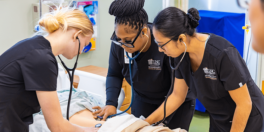 Three female UNC Pembroke nurses in black scrubs use stethoscopes to examine a patient in a hospital bed.