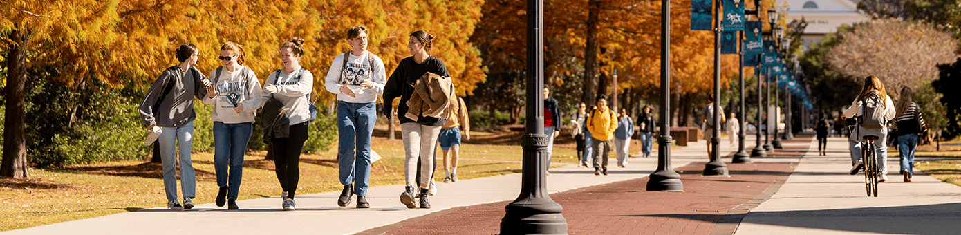 A college campus walkway during autumn with students walking and biking along a tree-lined path featuring orange and yellow foliage.
