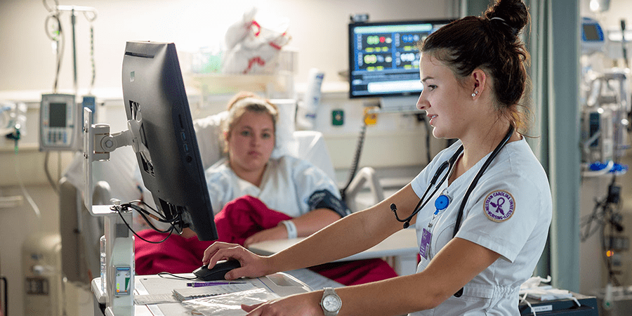 A female Western Carolina University nurse using a computer while attending to a patient lying in a hospital bed.
