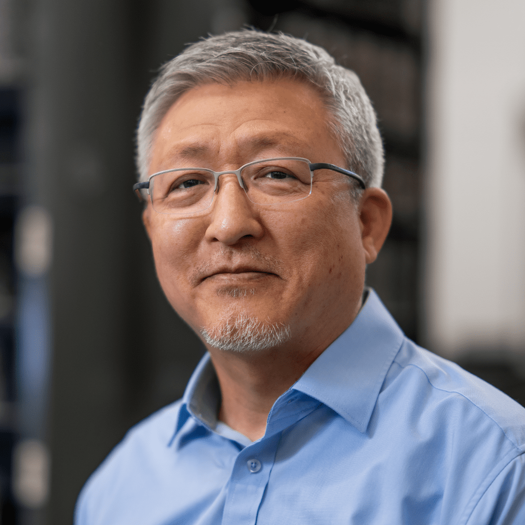 Dr. Richard Kim wearing a light blue button-up shirt and glasses, standing indoors with blurred shelves in background.