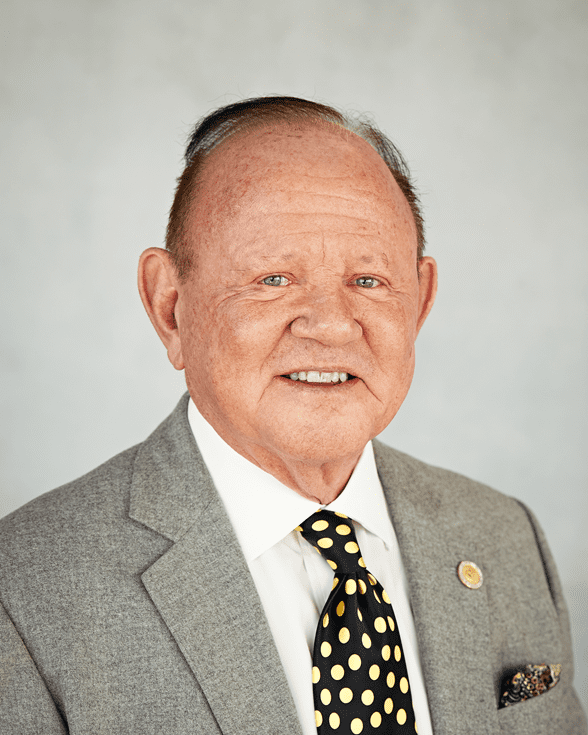 Headshot of Terry Hutchens wearing a gray suit jacket, white dress shirt, and black tie with yellow polka dots, standing against a light gray background.
