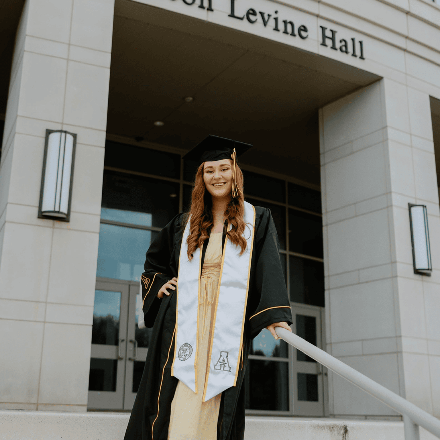 A female graduate standing on steps outside a building labeled "Levine Hall," wearing a black cap and gown with a white stole.
