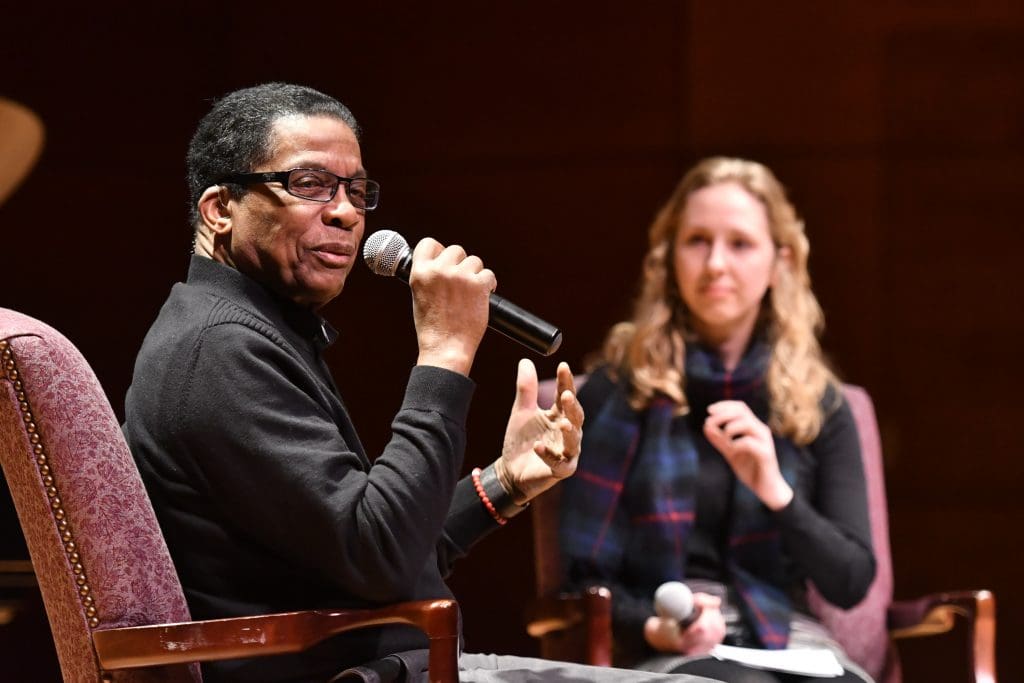 Musician speaks to audience while sitting in a chair