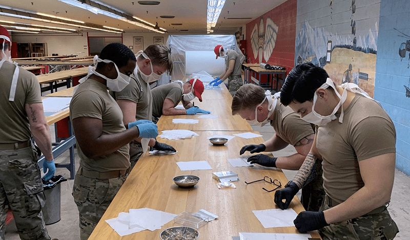 Soldiers stand around workshop tables assembling N95 masks