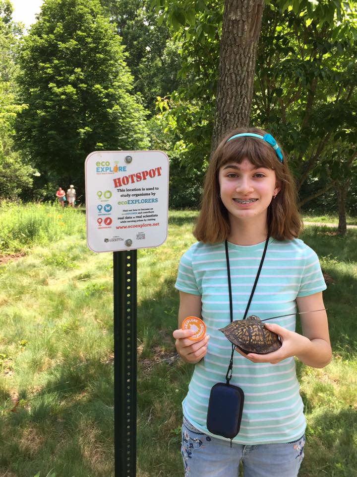 A girl holds a turtle while standing next to a "hot spot" sign
