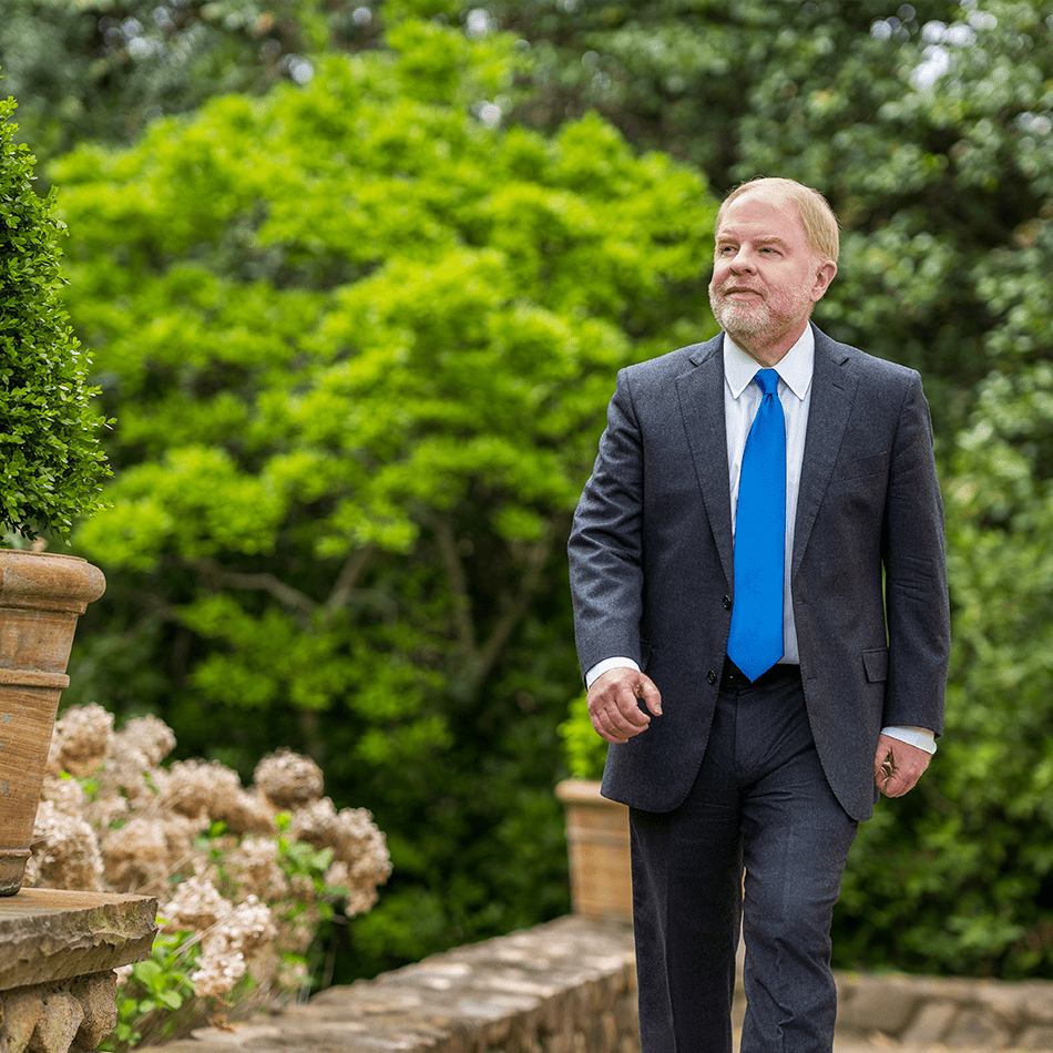 Peter Hans walking outdoors along a stone pathway surrounded by green foliage and potted plants.